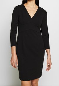 Musta vartalonmyötäinen mekko, jossa on pitkät hihat, V-kaula-aukko ja geometriset saumabroderiat. Valmistettu sileästä kankaasta, mekolla on virtaviivainen siluetti.