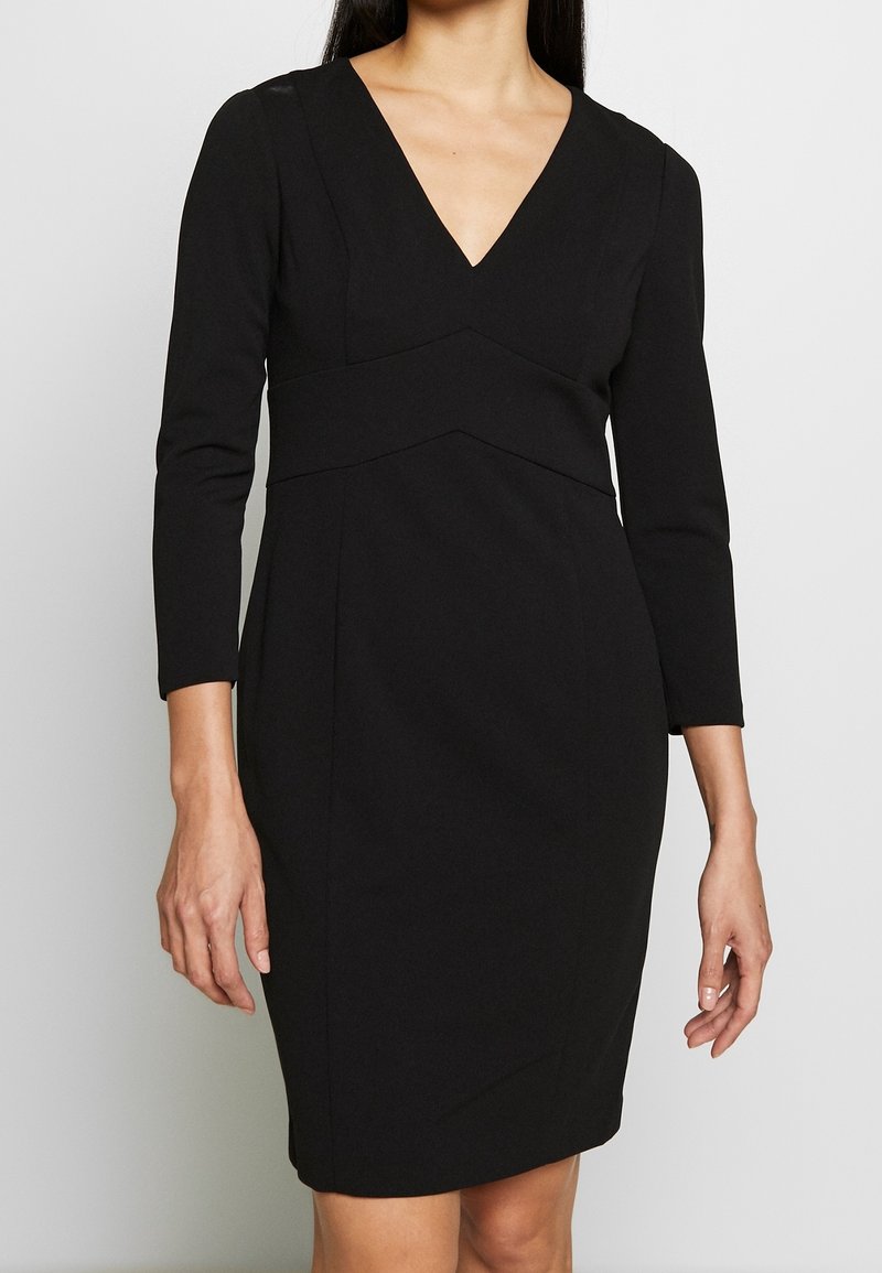 Musta vartalonmyötäinen mekko, jossa on pitkät hihat, V-kaula-aukko ja geometriset saumabroderiat. Valmistettu sileästä kankaasta, mekolla on virtaviivainen siluetti.