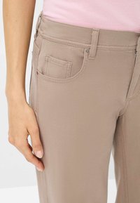Beige Denimhose mit glatter Textur, ausgestattet mit einer aufgesetzten Vordertasche und einem Reißverschluss. Der Stoff hat eine leichte Dehnbarkeit für hohen Tragekomfort.