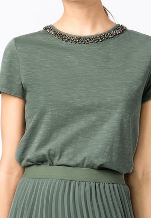 Femme portant un haut vert à manches courtes avec un col orné de perles, associé à une jupe plissée taille haute assortie.