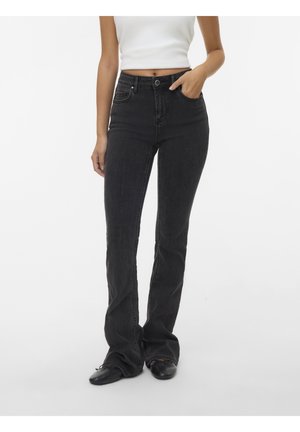 Kobieta ubrana w czarne jeansy z podwyższonym stanem i prostą nogawką, biały krótki top na ramiączkach oraz czarne baleriny, stojąca z jedną ręką w kieszeni.