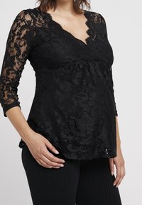 Top en dentelle noire à manches longues, avec un décolleté en V et un motif floral. Un tissu doux et texturé tombe délicatement sur la silhouette.