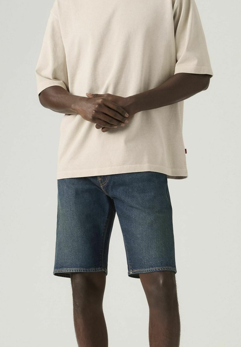 Homme portant une chemise beige à manches courtes et un short en denim bleu, debout les mains jointes devant un fond uni.
