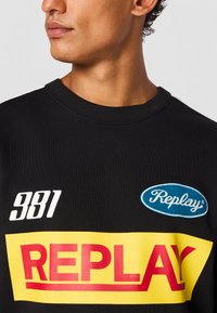 Svart sweatshirt med rund halsringning, stor röd "REPLAY"-text, blå oval logotyp och vit sifferdetalj "381". Mjuk tygkänsla.