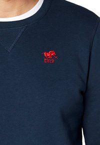 Granatowy sweter z okrągłym dekoltem, z czerwonym haftowanym logo konia oznaczonym napisem "POLO SYLT" na klatce piersiowej. Teksturowany materiał, dopasowany krój.
