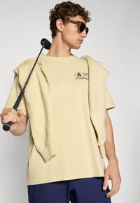 Light yellow cotton t-shirt with an embroidered design, paired with a light sweatshirt draped over the shoulders and dark blue shorts.