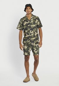 Camicia a maniche corte mimetica e pantaloni corti abbinati in verde e beige. Indossata con un cappello a secchiello nero e scarpe slip-on color cammello.