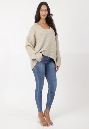 Suéter de punto beige con un escote amplio y dobladillo acanalado, combinado con jeans skinny de denim azul y sandalias de tacón transparente.