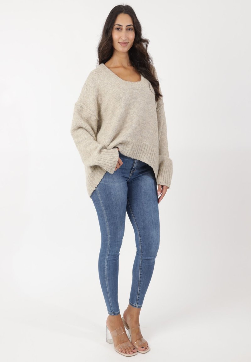Suéter de punto beige con un escote amplio y dobladillo acanalado, combinado con jeans skinny de denim azul y sandalias de tacón transparente.