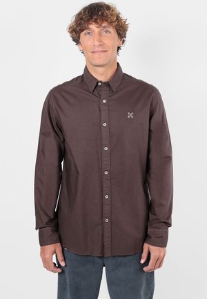 Homme aux cheveux bouclés portant une chemise marron foncé à manches longues boutonnée et un pantalon foncé, debout devant un fond blanc uni.