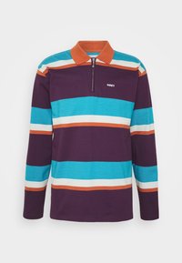 Fialová polo košile s dlouhým rukávem, zdobená horizontálními pruhy v té barvě, bílé a oranžové. Obsahuje zipový límec a bílé logo na hrudi.