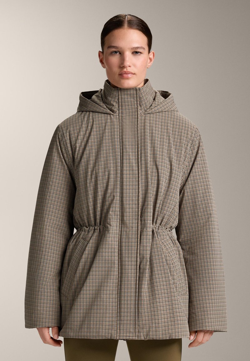 Vrouw draagt een geruite jas met kap, hoge kraag en getailleerde taille, staand tegen een eenvoudige neutrale achtergrond.