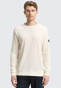 Crème-kleurig langslavend shirt van een gestructureerde stof, met een ronde hals en geribbelde manchetten, gecombineerd met donkere spijkerbroeken. Zwarte labeldetail op de mouw.