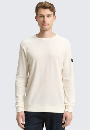 Crème-kleurig langslavend shirt van een gestructureerde stof, met een ronde hals en geribbelde manchetten, gecombineerd met donkere spijkerbroeken. Zwarte labeldetail op de mouw.