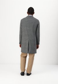 Grau karierter Mantel mit langem Schnitt, einem Schlitz hinten und schwarzen Knöpfen, kombiniert mit beigen Hosen und schwarzen Schuhen.