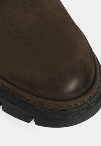 Botte en cuir marron avec une surface texturée, bout rond et semelle en caoutchouc noir. Présente des coutures visibles le long du bord et un talon surélevé.