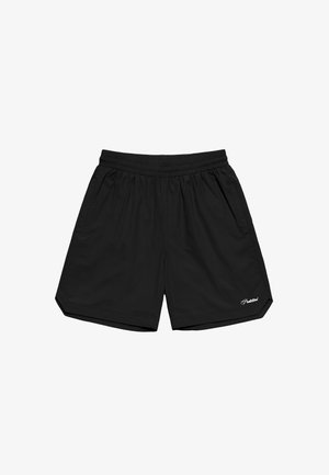 Inimene, kes kannab musti lühikesi pükse, valgeid sokke musta tekstiga ja pruune paeltega kingi, seisab ühevärvilise tausta ees.