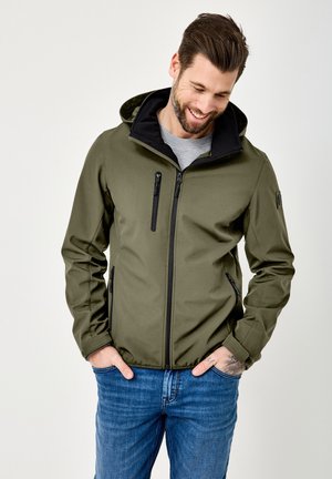 Lächelnder Mann mit Bart, der eine olivgrüne Kapuzenjacke über einem grauen Hemd und blauen Jeans trägt, die Hände in den Taschen, nach unten blickend.