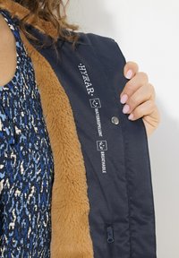 Marinblå vattentät och andningsbar jacka med en fawn-färgad fuskpälsfoder. Har präglade logotyper och en blixtlåsficka på sidan.