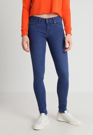 Person iført blå skinny jeans, hvide sneakers og en orange cropped langærmet top står mod en ensfarvet baggrund.