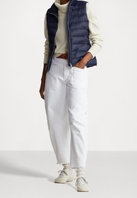 En person bär en marinblå puffväst över en krämfärgad tröja, vita vida byxor och ljusa sneakers, med händerna i fickorna.