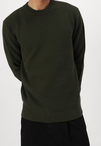 Olivově zelený svetr z texturovaného úpletu s kulatým výstřihem, žebrovanými manžetami a lemem; nošený s černými kalhotami, ruce překřížené za zády.