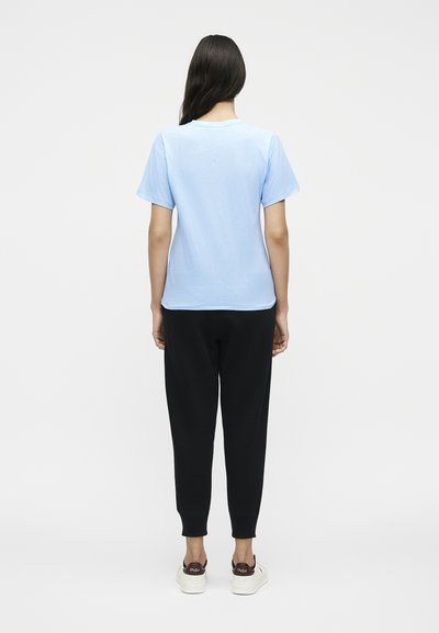 Γυναίκα με μακριά σκούρα μαλλιά, φοράει γαλάζιο κοντομάνικο πουκάμισο, μαύρο παντελόνι και λευκά αθλητικά παπούτσια, στέκεται με την πλάτη.