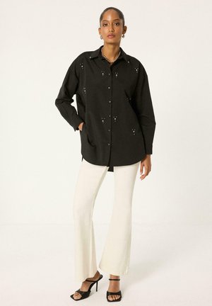 Femme portant une chemise noire oversize à boutons avec de petites découpes, pantalon blanc évasé et talons ouverts noirs, debout devant un fond uni.