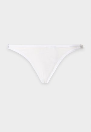 Kobieta stojąca boso ubrane w biały bralet Calvin Klein i czarne bieliznę w stylu bikini na jednolitym jasnym tle.