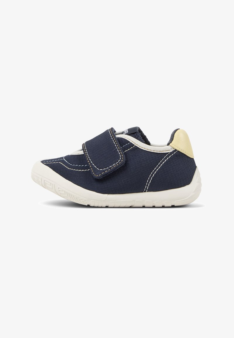 Chaussure en tissu bleu marine avec une bride en Velcro, semelle en caoutchouc blanche, tige texturée et accent beige au talon. Présente des détails de coutures visibles.
