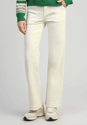 Pantalon crème droit avec poches avant, porté avec des baskets beiges et un pull vert rayé, personne debout avec une main dans la poche.