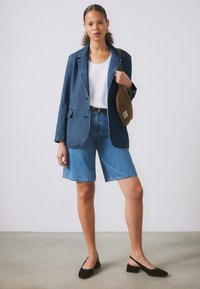 Blazer en denim bleu foncé, associé à un t-shirt blanc et un short en denim bleu clair. Des chaussures à talons carrés noires et un sac bandoulière marron complètent le look.