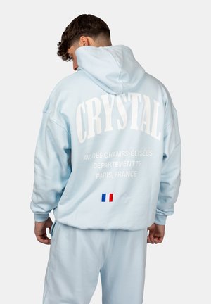 Lichtblauwe hoodie met de tekst "CRYSTAL" en locaties in Parijs op de achterkant, met een klein detail van de Franse vlag. Zachte textuur.