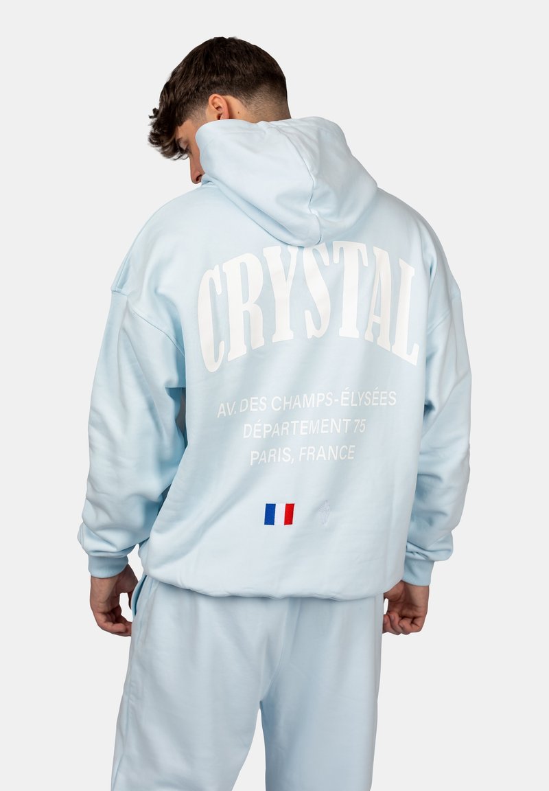 Sweat à capuche bleu clair avec "CRYSTAL" et des emplacements à Paris imprimés au dos, avec un petit détail du drapeau français. Texture douce.