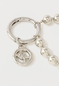 Hopeinen metallinen avaimenperä, jossa on sileä pyöreä rengas ja sarja pyöreitä helmiä. Mukana "MK"-logolla varustettu nimilappu. Kiillotettu pinta.
