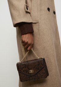Smeđa torbica sa uzorkom zmijske kože, zlatna lančana naramenica, pravokutnog oblika i zatvaranjem na preklop, drži se u ruci koja nosi smeđi pleteni rukav.