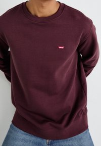 Burgundy sweatshirt med rund halsringning, mjukt bomullsmaterial och en röd Levi's-logotyp på bröstet. Kombineras med blå jeans.