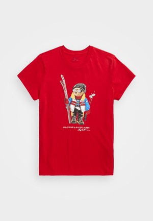 Czerwona bawełniana koszulka z grafiką przedstawiającą kreskówkowego niedźwiedzia w odzieży narciarskiej, trzymającego narty, z detalami graficznymi i logo marki na dole.