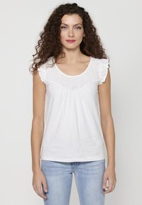 Top bianco senza maniche con dettagli in pizzo ricamato attorno al collo, vita arricciata e morbido materiale di cotone. Abbinato a jeans blu chiaro.
