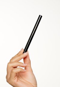 Zwarte oogpotlood eyeliner vastgehouden tussen vingers, slanke vorm, matte afwerking en geëmboste merknaam. Achtergrond is simpel wit.