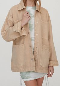 Veste en denim beige à coupe décontractée, quatre poches et détails de couture visibles. Comprend un col pointu et une fermeture par boutons pression.