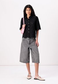 Svart øyelet skjorte med kort erm, kombinert med grå vide denimshorts og beige flate sko, accessorert med en rosa veske.