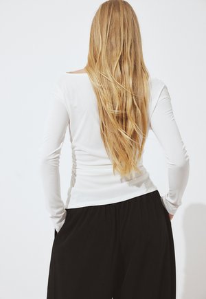 Žena s dlhými blond vlasmi, oblečená v bielej blúzke s dlhými rukávmi a čiernych nohaviciach, stojaca so založenými rukami vreckách, otočená chrbtom ku kamere.