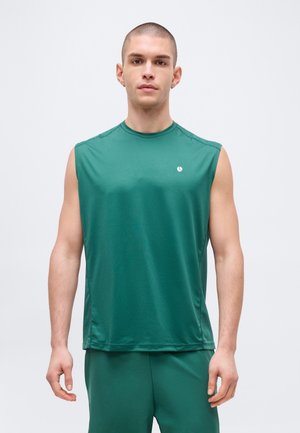 Άντρας με κοντά μαλλιά που φοράει πράσινο αμάνικο αθλητικό μπλουζάκι και αντίστοιχο σορτς, κοιτάζοντας μπροστά πάνω σε απλό φωτεινό φόντο.