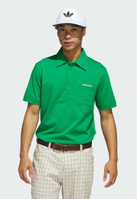 Grön polo med knappslå och bröstficka till vänster. Stylar med krämfärgade rutiga byxor och en vit skärmfri hatt med en svart logotyp.