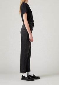 Femme debout en t-shirt noir, jean noir taille haute, chaussettes blanches et mocassins noirs sur fond gris clair, vue de profil.