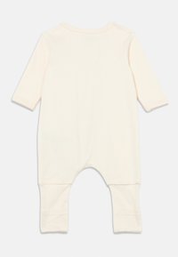 Kremfarget langermet heldrakt med rundet halslinning, mansjetter ved håndledd og ankler, og en løs passform. Glatt, myk stofftekstur.