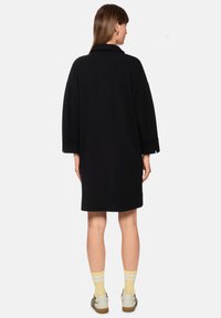 Schwarzer, übergroßer Mantel mit niedrigem Kragen, überschnittenen Schultern und langen Ärmeln. Verfügt über einen geraden Schnitt und einen knieumspielenden Saum.