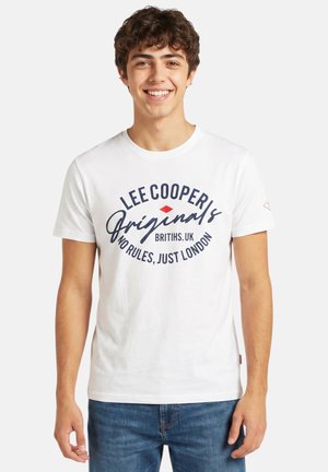 Jeune homme souriant portant un t-shirt graphique blanc Lee Cooper et un jean bleu, debout devant un fond blanc uni.