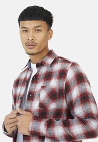 Młody mężczyzna z krótkimi czarnymi włosami, ubrany w koszulę w kratę w kolorach czerwonym, białym i szarym, noszącą białą koszulkę pod nią, na jednolitym tle.